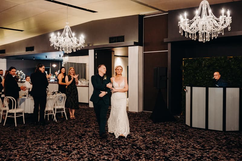 Bride Madison and groom Brendon walk arm in arm into the reception room at Sandstone Point Hotel — Pumicestone Room while guests applaud and a DJ stands behind a booth.