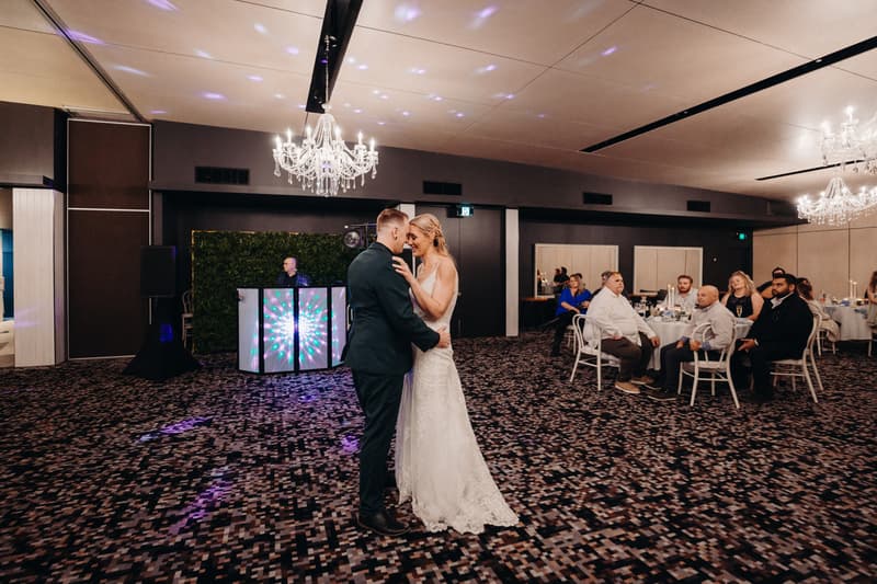 Bride Madison and groom Brendon share a dance on the carpeted floor of the Pumicestone Room at Sandstone Point Hotel, with seated guests watching and a DJ booth illuminated in the background.