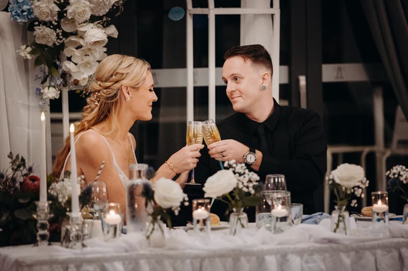 Bride Madison and groom Brendon toast with champagne glasses at their reception table decorated with white roses and candles in the Pumicestone Room at Sandstone Point Hotel.