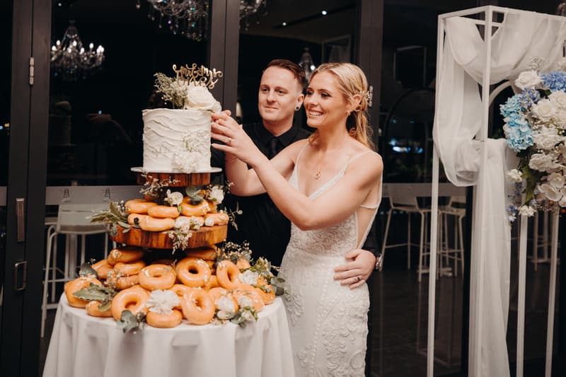 Bride Madison and groom Brendon cut a wedding cake topped with a 'Mr & Mrs' sign at Sandstone Point Hotel — Pumicestone Room, surrounded by a tiered display of donuts on a round table.