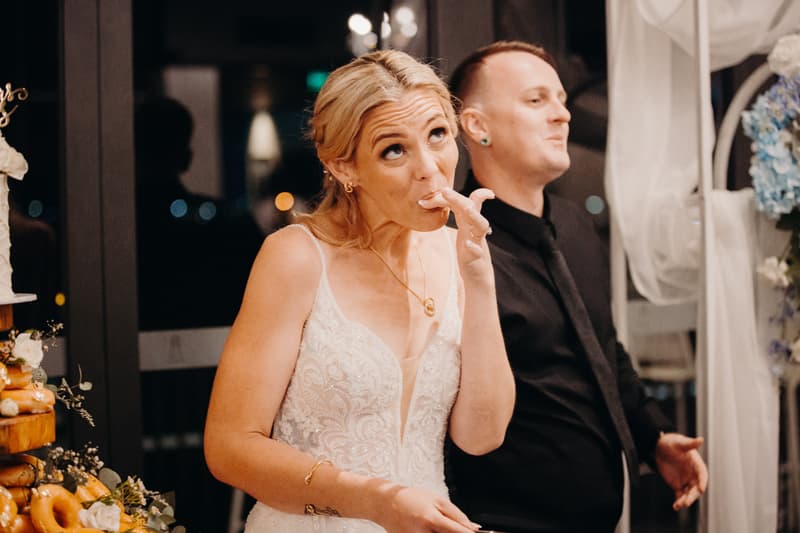 The bride Madison in a white lace wedding dress is licking her finger while standing next to the groom Brendon dressed in a black shirt and tie at the Sandstone Point Hotel — Pumicestone Room during the reception stage.