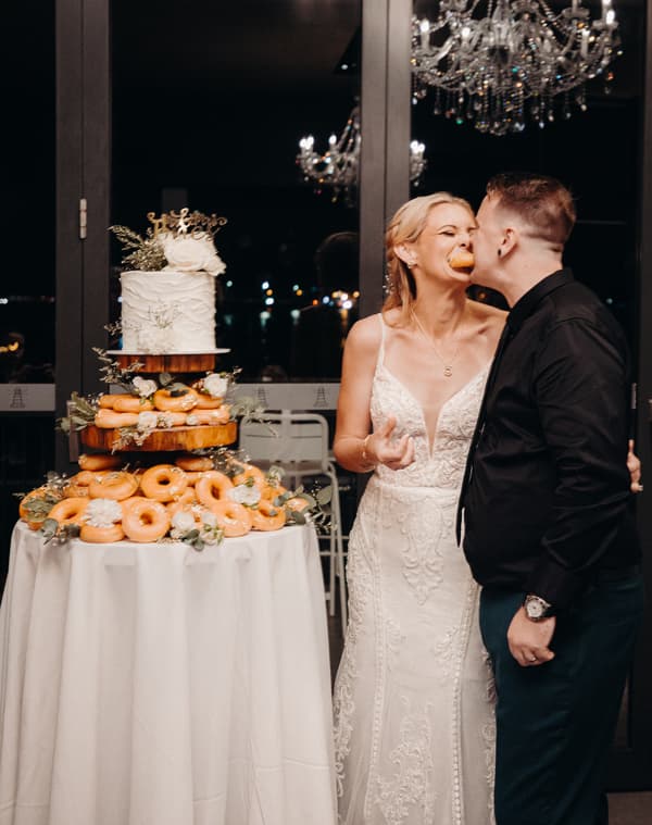 The bride Madison and groom Brendon stand next to a wedding cake and a large display of donuts at Sandstone Point Hotel — Pumicestone Room. Madison has a donut in her mouth while Brendon leans in close to her.