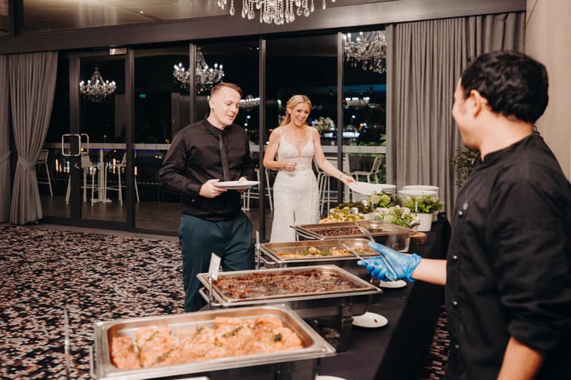 The bride Madison and groom Brendon are at the buffet table in the Pumicestone Room at Sandstone Point Hotel, selecting food served by a catering staff member.