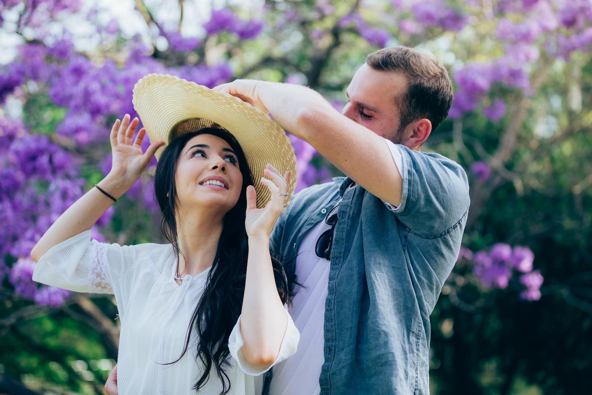 The couple shares a playful moment with a straw hat under vibrant purple jacaranda trees.