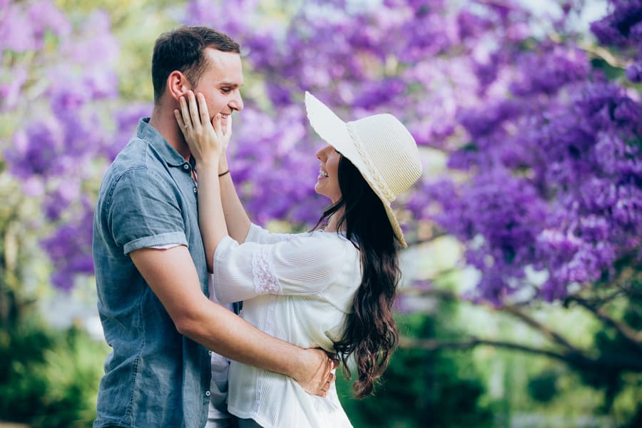 The couple embraces warmly, surrounded by blooming purple jacaranda trees at UQ Lakes.