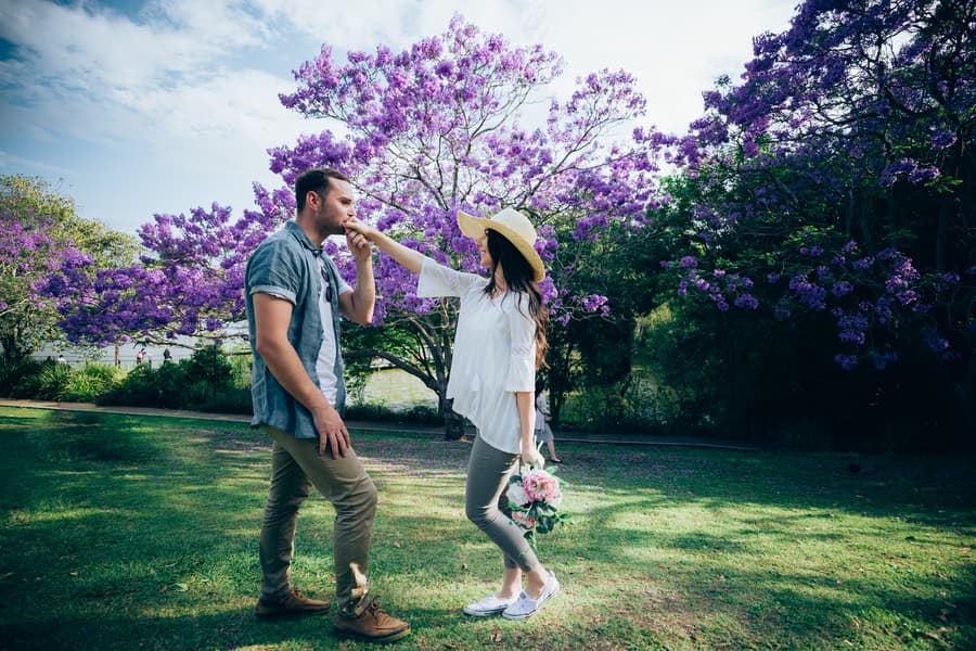 The couple stands on grass, exchanging a tender look, with a bouquet in hand and jacarandas in the background.