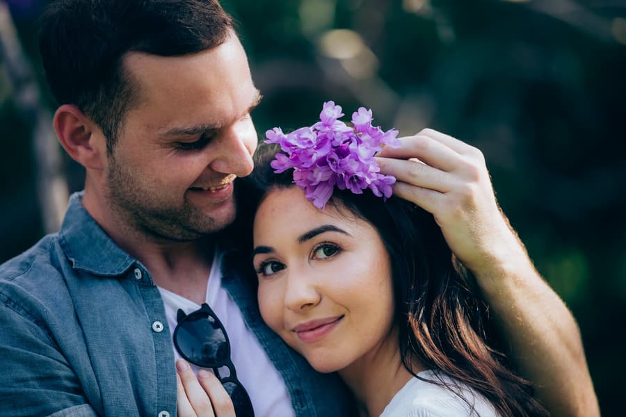 The couple shares a close moment, with one placing a purple flower in the other's hair, smiling softly.