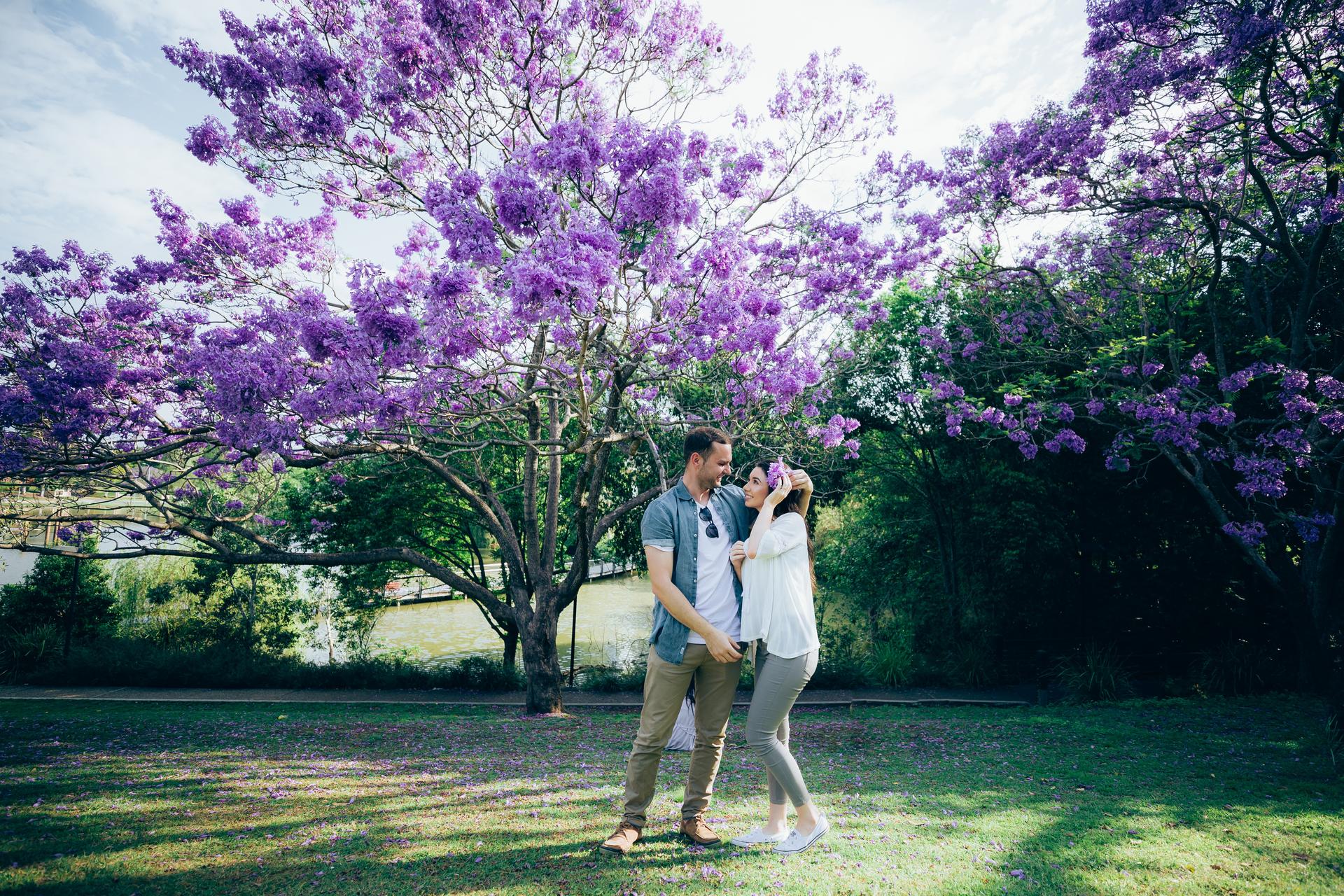 The couple stands together under a jacaranda tree, sharing a joyful laugh in the sunlight.