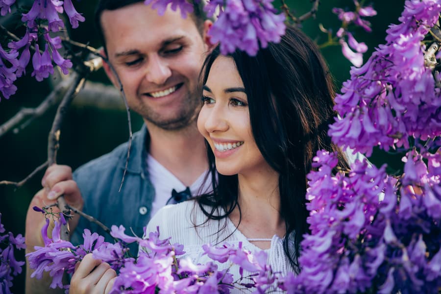 The couple is surrounded by purple jacaranda blossoms, smiling and enjoying each other's company.