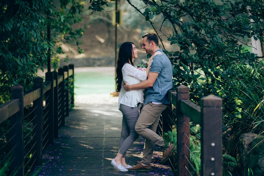 The couple stands on a wooden bridge, gazing into each other's eyes, surrounded by greenery.