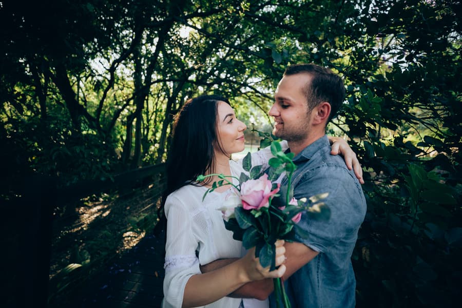 The couple stands close, holding a bouquet, with lush foliage in the background, sharing a tender gaze.