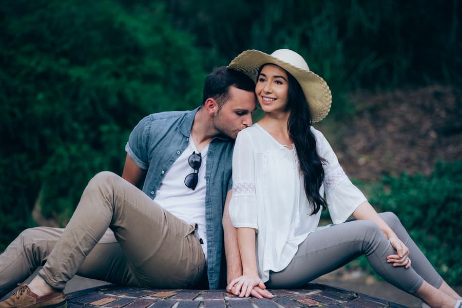 The couple sits together on a brick surface, one leaning in for a kiss on the cheek, both relaxed.