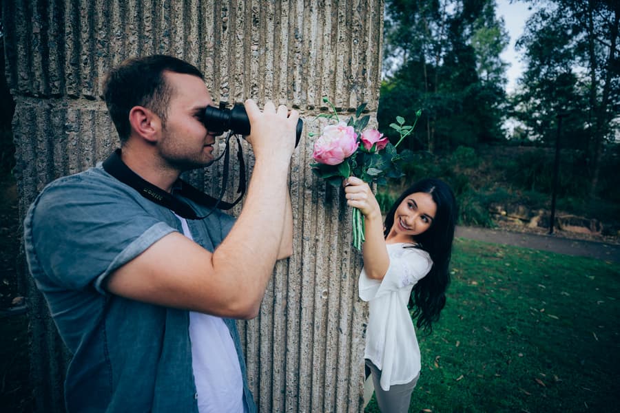 One of the couple playfully holds a bouquet while the other takes a photo with a camera, both smiling.