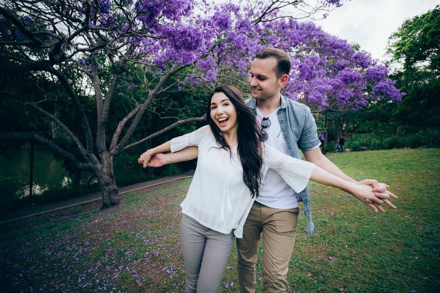 The couple joyfully poses under a blooming jacaranda tree at UQ Lakes, with arms outstretched.