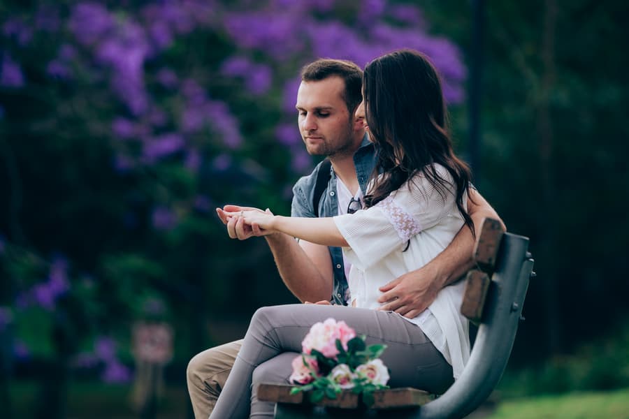 The couple sits closely on a bench at UQ Lakes, examining a ring, with a bouquet beside them.