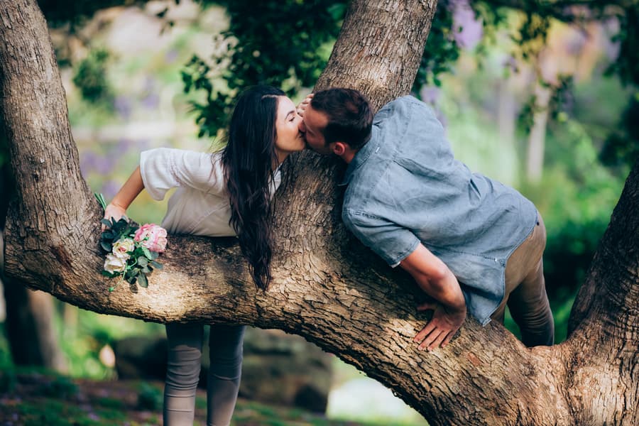 The couple shares a kiss while playfully leaning on a tree branch at UQ Lakes, holding a bouquet.
