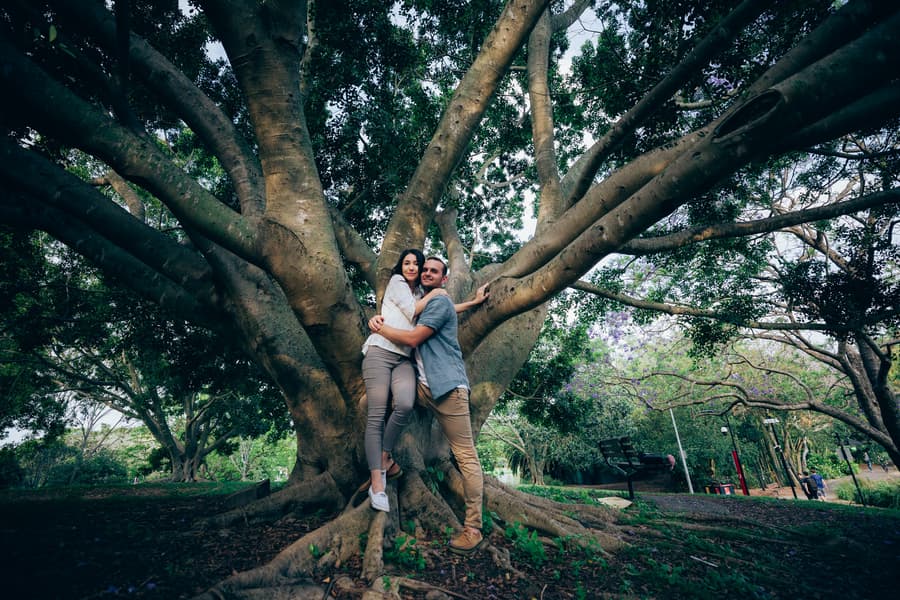 The couple embraces while standing among the large roots of a tree at UQ Lakes, surrounded by greenery.