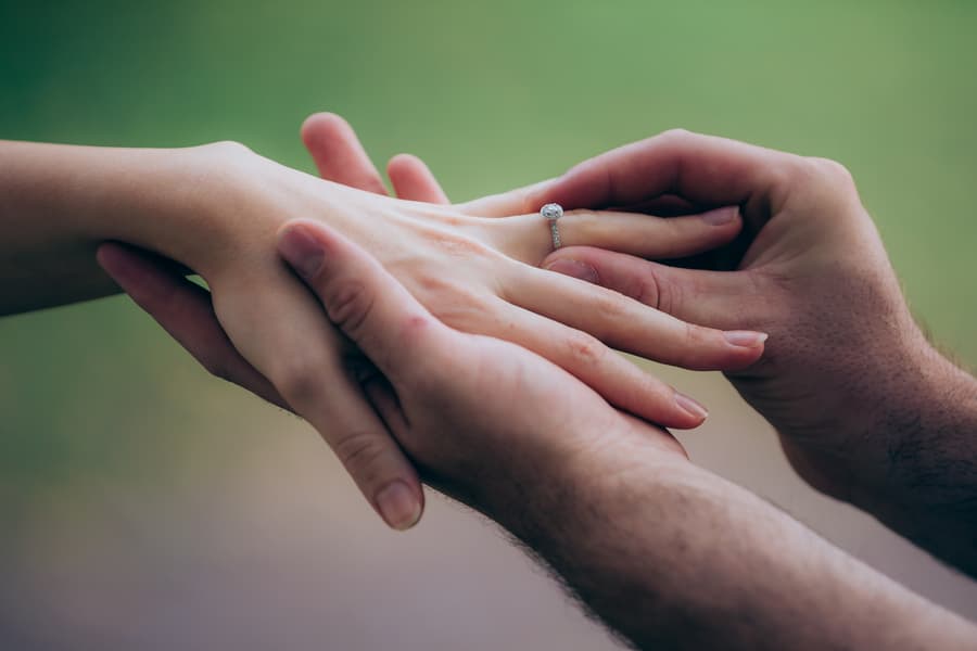 Close-up of hands showing an engagement ring, with soft focus on the background.
