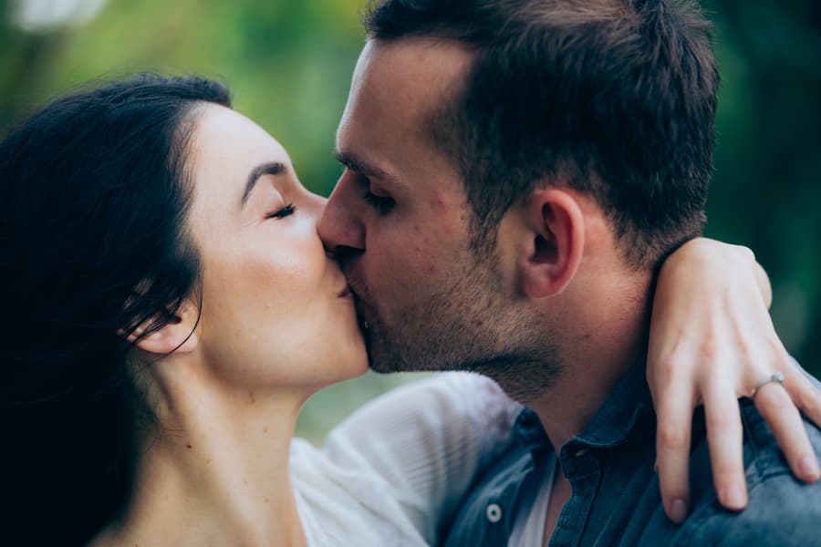 The couple shares a kiss, with one wearing an engagement ring, against a natural backdrop.