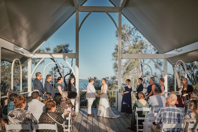 Brides Mandy and Selina hold hands at the altar during their wedding ceremony at Sandstone Point Hotel — The Pavilion, surrounded by their wedding party and seated guests.