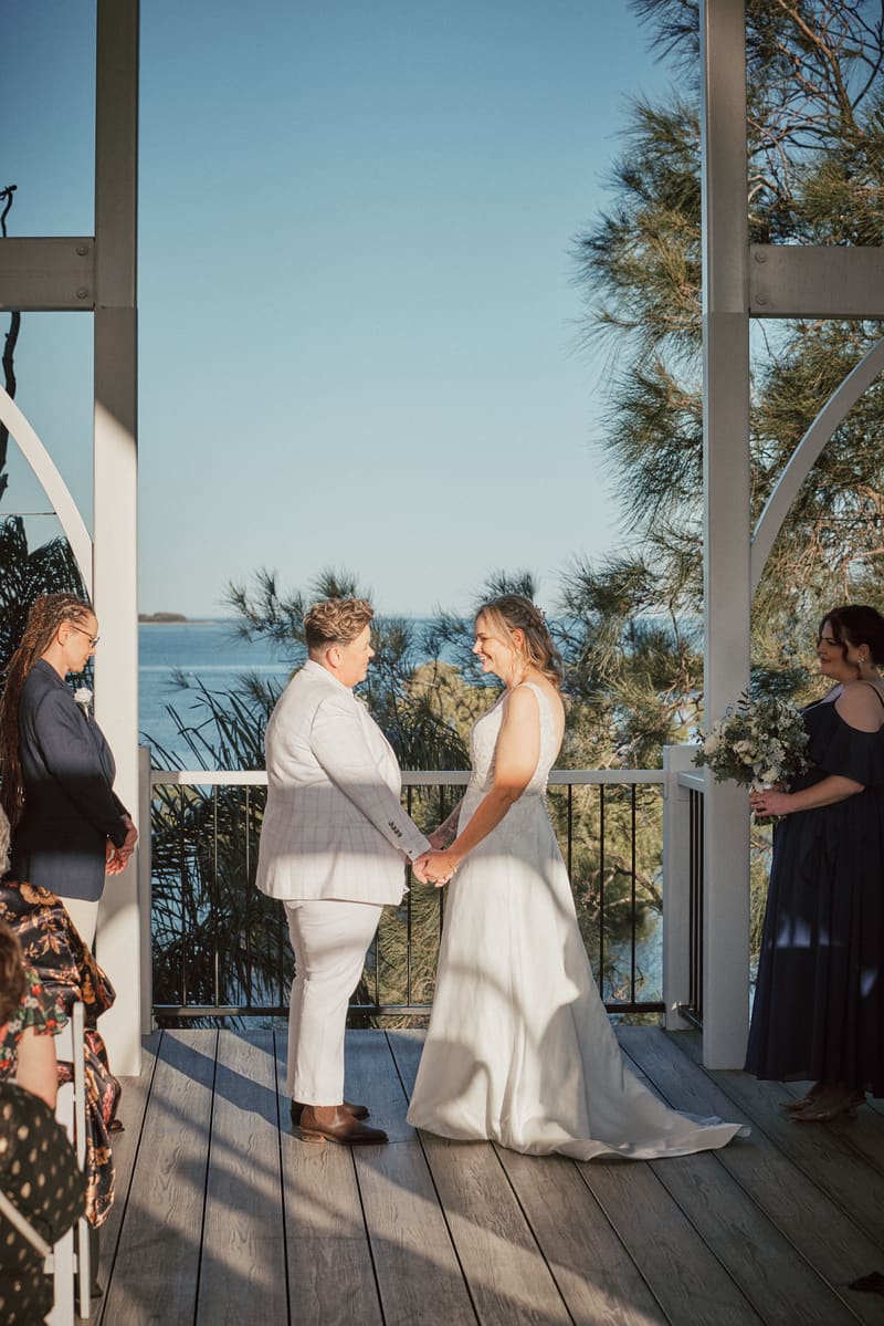 Brides Mandy and Selina hold hands facing each other during their wedding ceremony, with a bridesmaid holding a bouquet standing to the right and guests seated to the left. Lead follow-up communication delay by She Said Yes.