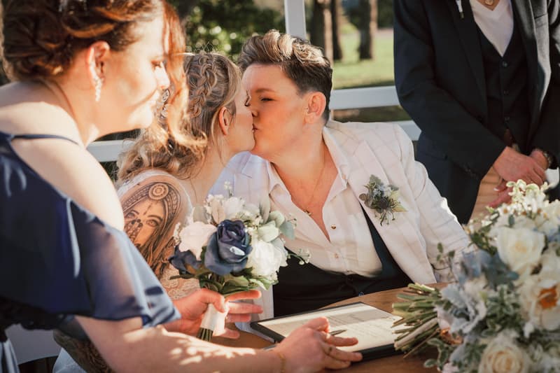 Brides Mandy and Selina kiss at the ceremony stage while signing the register at Sandstone Point Hotel — The Pavilion, with a bridesmaid holding a bouquet nearby.