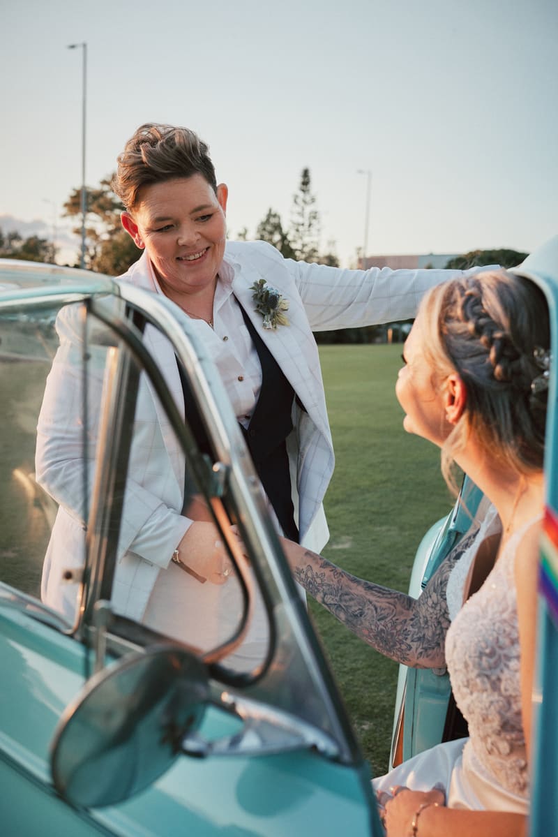 Bride Selina in a white suit with a boutonniere smiles while holding hands with bride Mandy, who is seated in a vintage turquoise car wearing a lace wedding dress with visible tattoos on her arm,. Best wedding photographer Moreton Bay by She Said Yes.