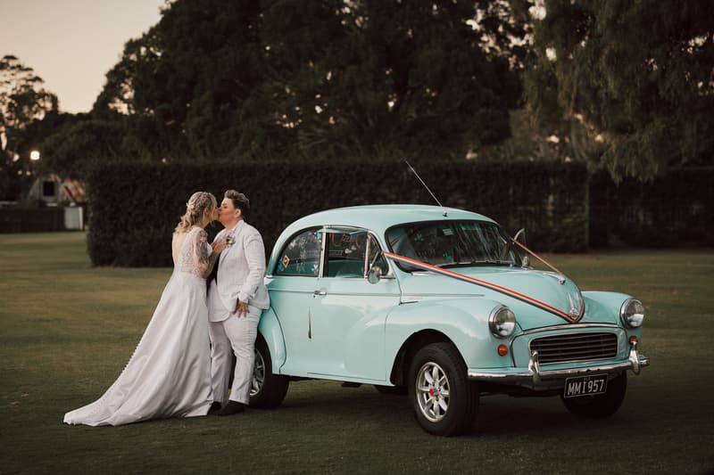 Brides Mandy and Selina kiss beside a vintage light blue car decorated with ribbons on the lawn at Sandstone Point Hotel.
