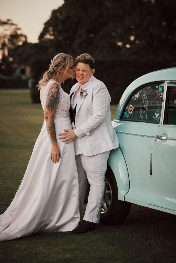 Mandy in a white wedding gown kisses Selina, who is wearing a white suit, on the cheek while leaning against a light blue vintage car at Sandstone Point Hotel.