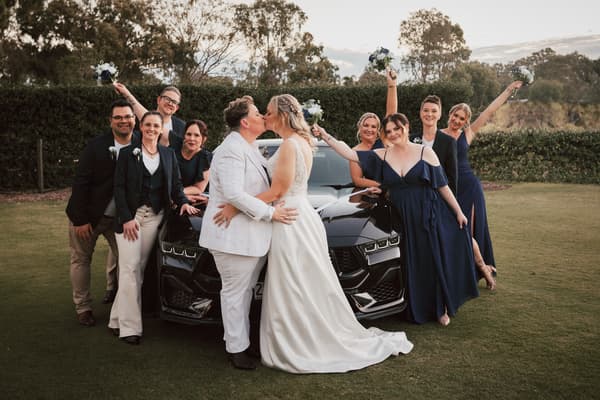 Brides Mandy and Selina kiss in front of a black car, surrounded by their wedding party dressed in navy and dark suits, some holding bouquets. May wedding sunset Brisbane by She Said Yes.