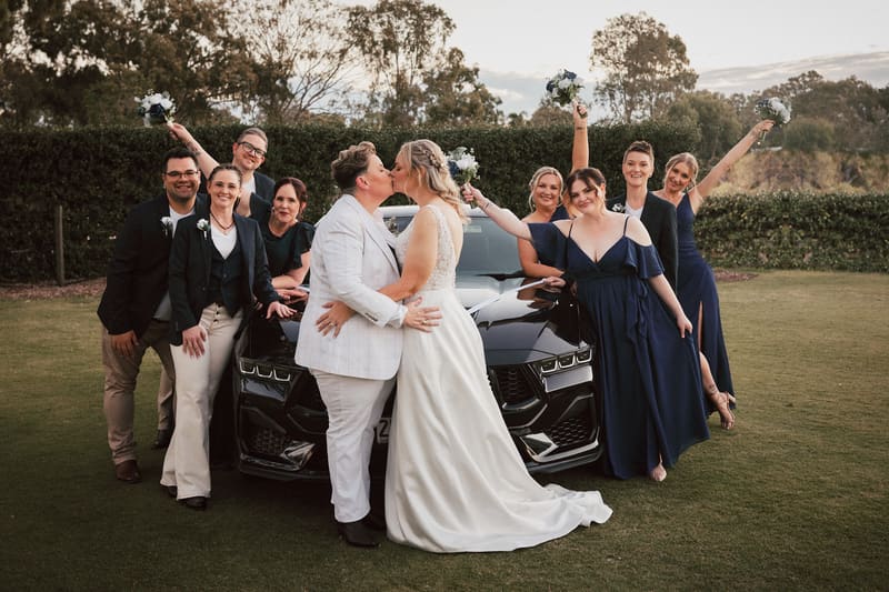 Brides Mandy and Selina kiss in front of a black car, surrounded by their wedding party dressed in navy and dark suits, some holding bouquets. Engagement parties by She Said Yes.