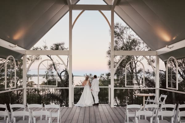 Brides Mandy and Selina stand side by side on a wooden deck, facing a scenic view of trees and water during sunset, with empty white chairs arranged around them. Wedding family photos by She Said Yes.