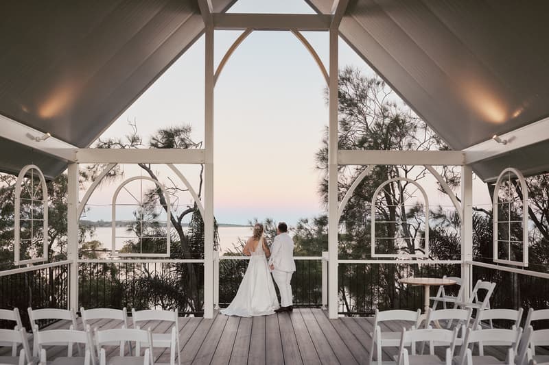 Brides Mandy and Selina stand side by side on a wooden deck at Sandstone Point Hotel, facing a scenic view of trees and water during sunset, with empty white chairs arranged around them.
