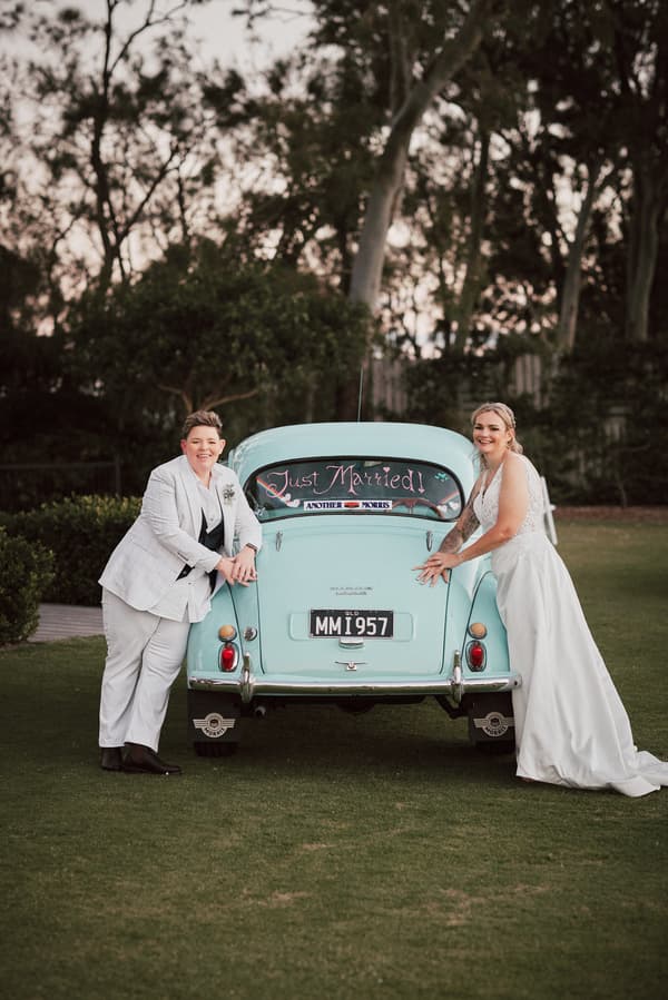Brides Mandy and Selina pose beside a vintage light blue car with a 'Just Married!' sign on the rear window at Sandstone Point Hotel.