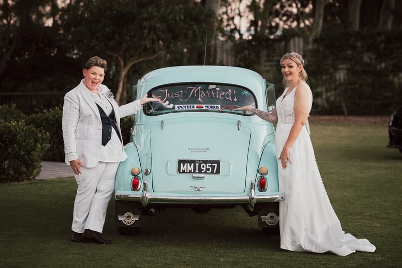 Brides Mandy and Selina stand on either side of a light blue vintage car with a 'Just Married!' sign on the rear window at Sandstone Point Hotel.