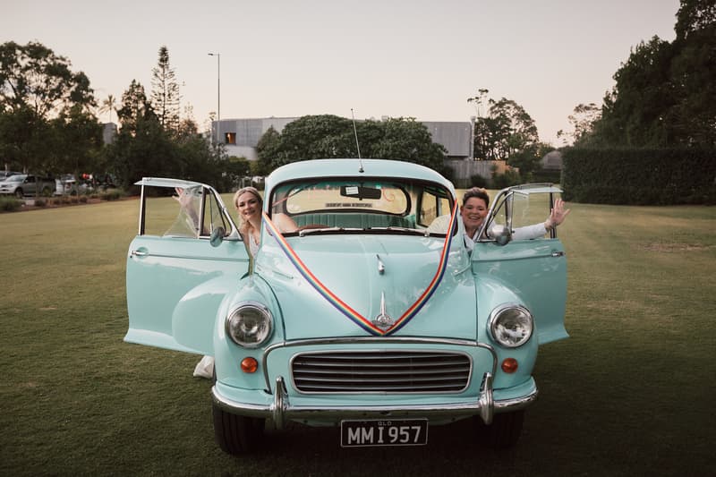 Brides Mandy and Selina pose with open doors of a vintage light blue car decorated with a rainbow ribbon at Sandstone Point Hotel on a grassy lawn.