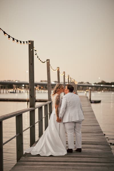 Brides Mandy and Selina share a kiss while holding hands on a wooden pier decorated with string lights. Destination wedding photographer by She Said Yes.