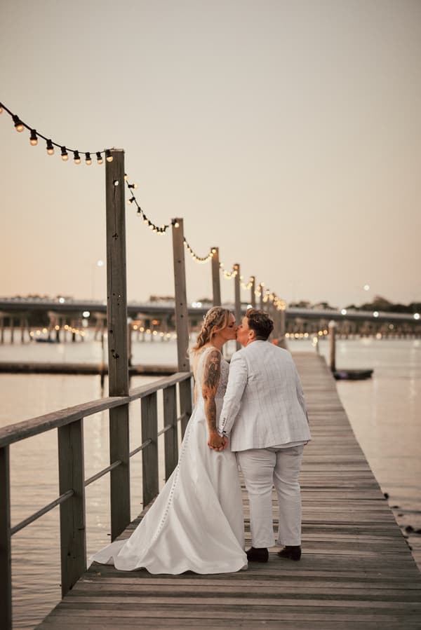 Brides Mandy and Selina share a kiss while holding hands on a wooden pier decorated with string lights at Sandstone Point Hotel during their couple portraits session.