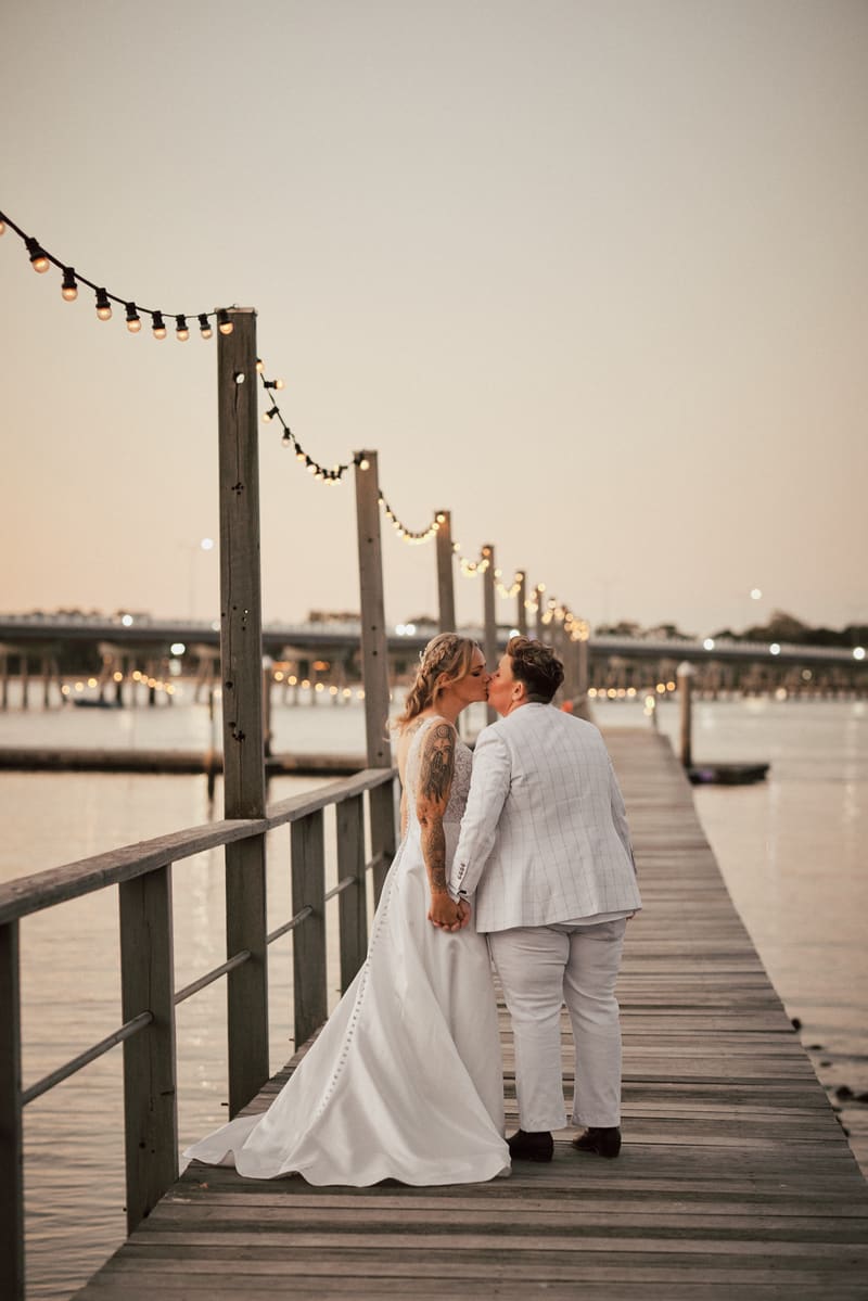 Brides Mandy and Selina share a kiss while holding hands on a wooden pier decorated with string lights. Wedding photographer Brisbane by She Said Yes.