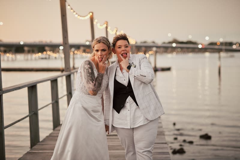 Brides Mandy and Selina pose playfully on a wooden pier at Sandstone Point Hotel during their couple portraits session, with string lights and water in the background at dusk.