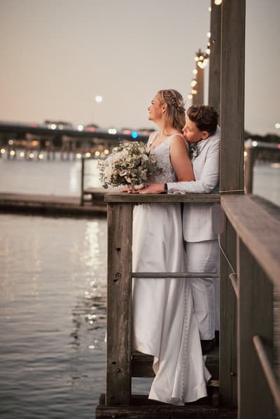 Brides Mandy and Selina embrace on a wooden pier, with Mandy holding a bouquet and Selina leaning in close behind her, overlooking the water at dusk. Wedding photography contracts by She Said Yes.
