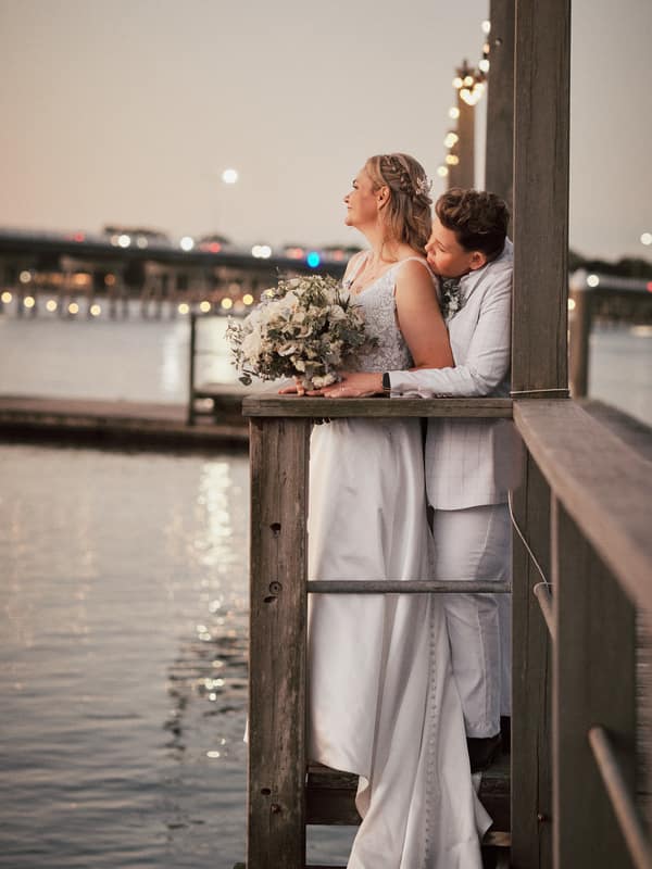 Brides Mandy and Selina embrace on a wooden pier at Sandstone Point Hotel, with Mandy holding a bouquet and Selina leaning in close behind her, overlooking the water at dusk.