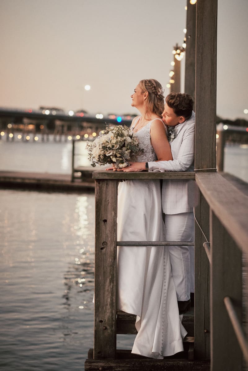 Brides Mandy and Selina embrace on a wooden pier, with Mandy holding a bouquet and Selina leaning in close behind her, overlooking the water at dusk. ABIA 2018 finalist wedding photography by She Said Yes.