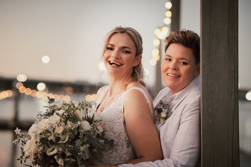 Brides Mandy and Selina pose together at Sandstone Point Hotel during their couple portraits session, with Mandy holding a large bouquet of white and green flowers and Selina standing behind her, both smiling.
