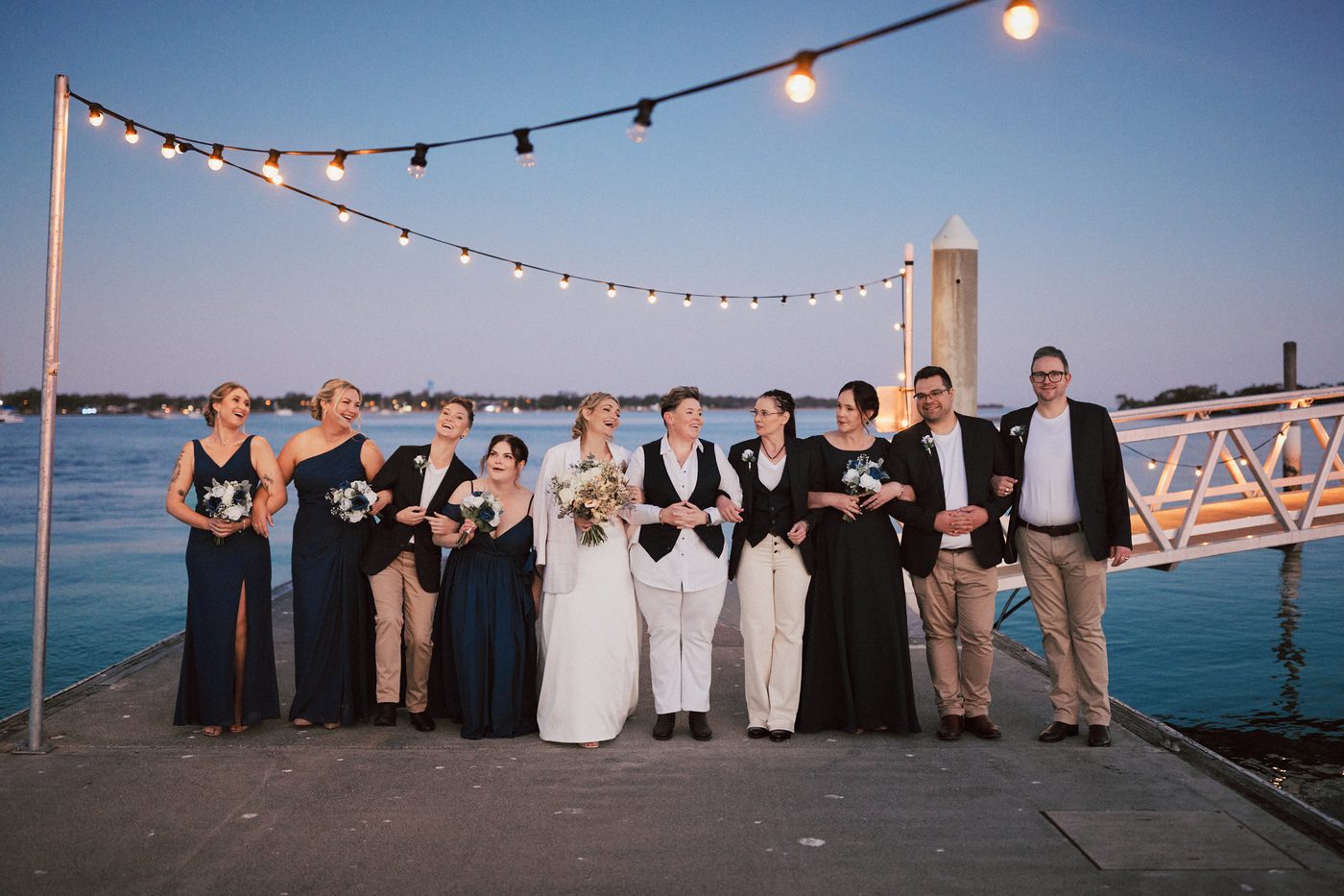 Mandy and Selina stand arm in arm with their wedding party on a pier, with string lights overhead and water in the background. Cinematic wedding photos by She Said Yes.