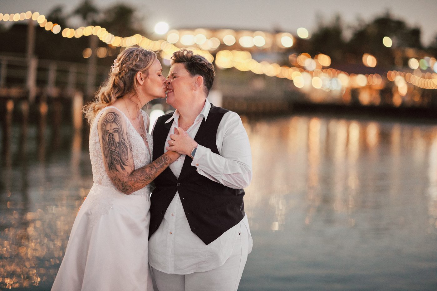 Brides Mandy and Selina share a close moment by the water, with string lights softly glowing in the background at dusk. Lead follow-up communication issues by She Said Yes.