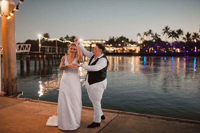 Brides Mandy and Selina dance together on a waterfront promenade, with string lights and palm trees visible across the water. Engagement ceremony by She Said Yes.