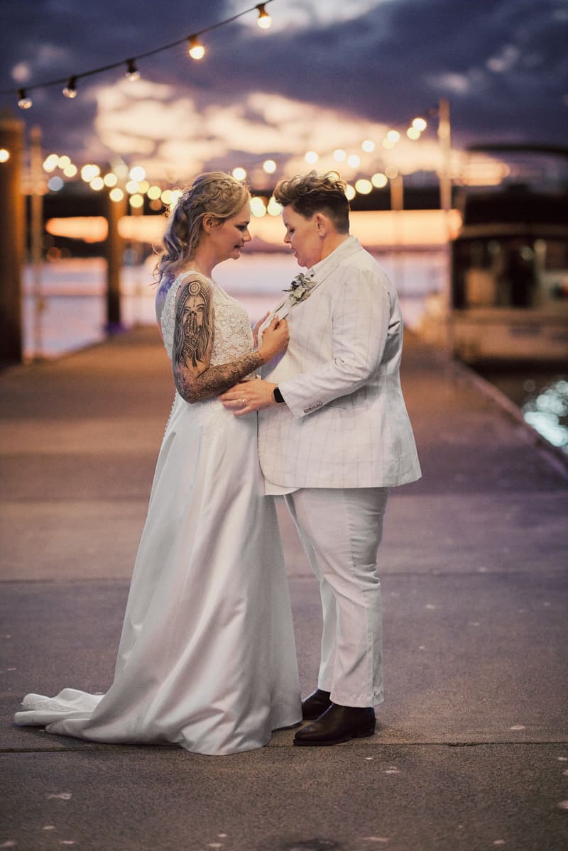 Brides Mandy and Selina stand facing each other on a pier, with string lights overhead and a boat docked in the background. Cinematic photography wedding by She Said Yes.