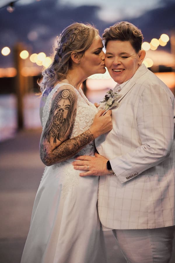 Bride Mandy kisses bride Selina on the cheek during their couple portraits at Sandstone Point Hotel, with Mandy wearing a white lace wedding dress and Selina in a light-colored checkered suit.