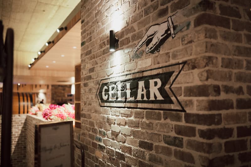 Interior view of the Sandstone Point Hotel — The Cellar showing a brick wall with a painted sign reading 'CELLAR' and a pointing hand above it, with blurred floral arrangements and wooden barrels in the background.